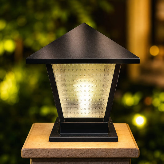 A black outdoor solar post cap light with a textured glass panel, glowing with warm yellow light, mounted on a stone post with a blurred garden background.