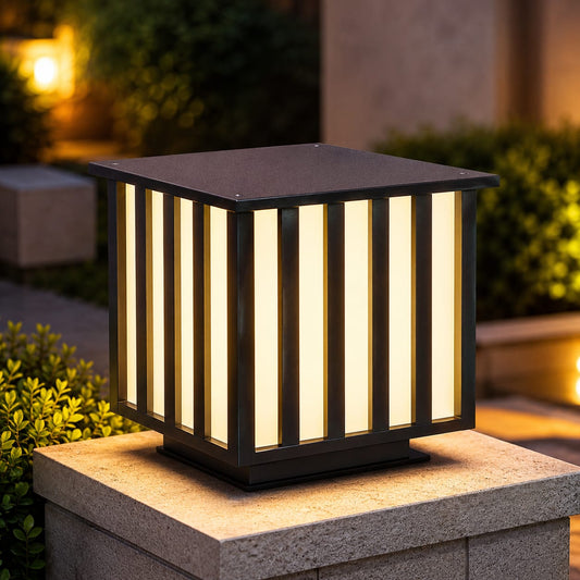 A modern square outdoor solar post cap light with vertical slats glowing with warm yellow light, mounted on a stone pillar in a garden setting.